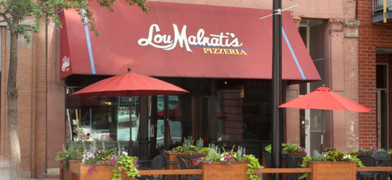 Cozy downtown pizzeria storefront with red awning, matching patio umbrellas, wooden planters of flowers and outdoor seating.