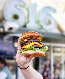 Close-up of a hand holding a juicy double bacon cheeseburger with melted cheddar, lettuce and brioche bun in front of a blurred street-side restaurant sign.