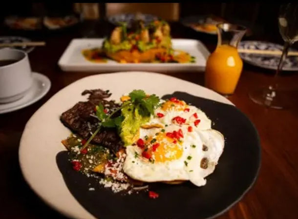 Vibrant steak-and-eggs brunch: two sunny-side-up eggs with chopped red pepper, smashed avocado, seared steak and roasted veggies on a black-and-white plate, with coffee cup and small carafe of orange juice on a wooden restaurant table.