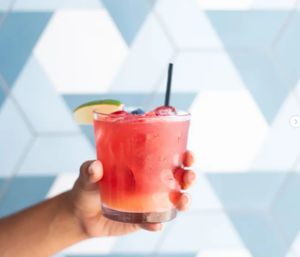 Hand holding a pink berry cocktail in a short glass with ice, black straw, lime wedge and berry garnish against a blue geometric tile background