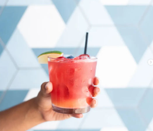 Hand holding a pink berry cocktail in a short glass with ice, black straw, lime wedge and berry garnish against a blue geometric tile background