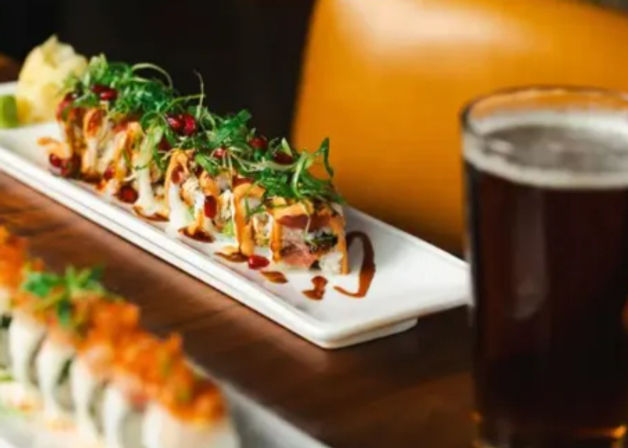 Colorful sushi rolls topped with microgreens and drizzled sauces on white rectangular plates beside a pint of dark beer on a wooden restaurant table.
