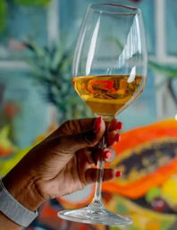 Hand with red-painted nails holding a wine glass filled with golden wine against a colorful tropical backdrop featuring papaya and leafy plants.