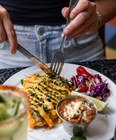 Hands cutting into a breaded fillet drizzled with spicy orange sauce and chopped greens on a white plate, accompanied by purple cabbage slaw, a lime wedge and a small cup of pinto beans topped with cheese.