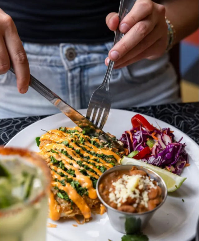 Hands cutting into a breaded fillet drizzled with spicy orange sauce and chopped greens on a white plate, accompanied by purple cabbage slaw, a lime wedge and a small cup of pinto beans topped with cheese.