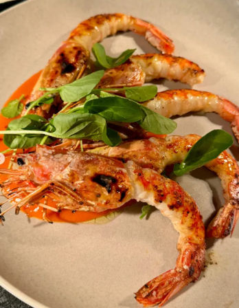 Charred jumbo prawns (shrimp) drizzled with red pepper sauce and topped with fresh microgreens on a stone plate — succulent grilled seafood dish.