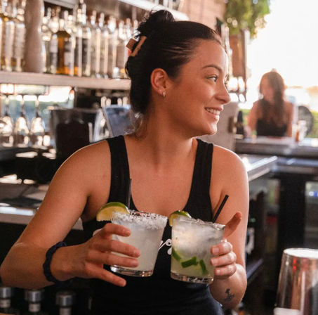 Smiling bartender carrying two frosty lime margaritas with salted rims and lime wedges at a bustling bar counter