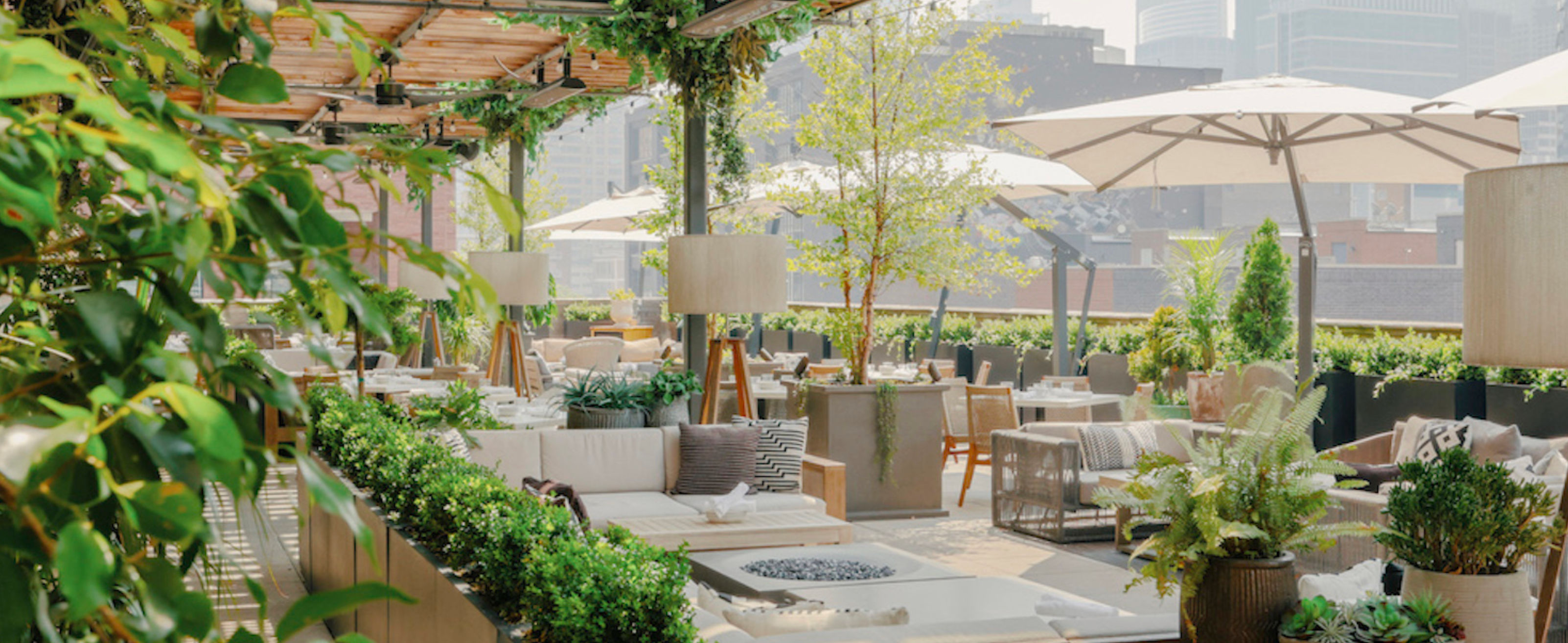 Lush urban rooftop terrace with cozy cushioned lounges, dining tables under umbrellas, potted plants and a pergola overlooking the city skyline