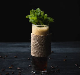 Tall cold brew coffee in a highball glass wrapped in burlap, frothy top crowned with fresh mint sprigs, coffee beans scattered on dark wood against a black background.