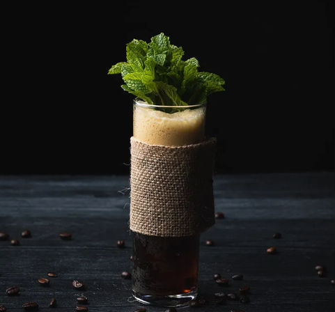 Tall cold brew coffee in a highball glass wrapped in burlap, frothy top crowned with fresh mint sprigs, coffee beans scattered on dark wood against a black background.