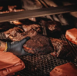 Gloved hand lifting a bark-crusted smoked brisket from a hot barbecue smoker rack, wrapped sides visible