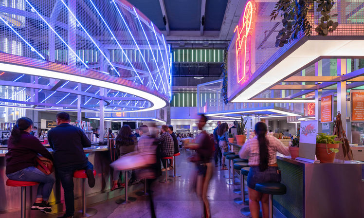 Vibrant downtown indoor food hall at night with circular blue and pink LED canopy, neon signs, bar stools, potted plants and bustling diners