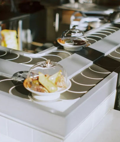 Conveyor-belt sushi-style service with small white plates of bite-sized appetizers covered under clear domes gliding along a rotating track in a modern restaurant