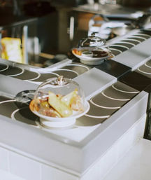 Conveyor-belt sushi-style service with small white plates of bite-sized appetizers covered under clear domes gliding along a rotating track in a modern restaurant