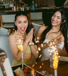 Two women celebrating at a restaurant table with dessert sparklers—smiling, clapping, festive dining moment.