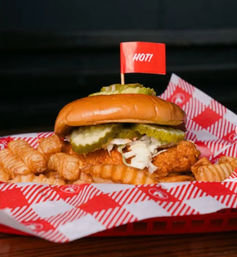 Spicy crispy fried chicken sandwich with pickles and shredded slaw on a brioche bun, served with crinkle-cut fries in a red checkered diner basket and topped with a small red "HOT!" flag