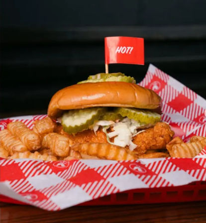 Spicy crispy fried chicken sandwich with pickles and shredded slaw on a brioche bun, served with crinkle-cut fries in a red checkered diner basket and topped with a small red "HOT!" flag