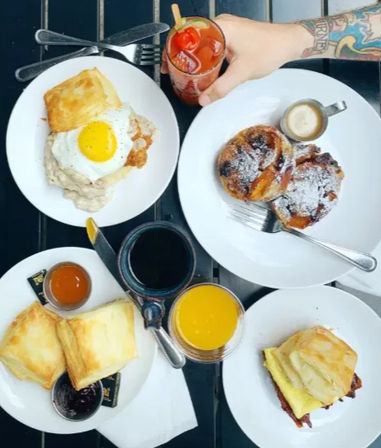 Outdoor patio brunch spread: biscuits with gravy and egg, jam-topped pastries, cinnamon rolls dusted with powdered sugar, coffee, orange juice and a Bloody Mary held by a tattooed arm.