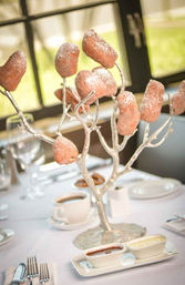 Silver branch dessert centerpiece holding sugar-dusted mini doughnuts like ornaments on a white event table with coffee cups, dipping sauces, plates and polished cutlery.