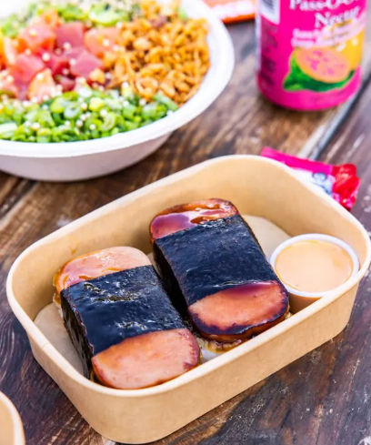 Two Hawaiian Spam musubi in a compostable takeout tray — grilled Spam slices on rice wrapped with nori, small dipping sauce cup, with a poke bowl and canned drink blurred on a wooden table.