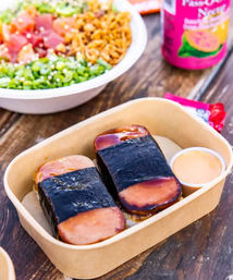 Two Hawaiian Spam musubi in a compostable takeout tray — grilled Spam slices on rice wrapped with nori, small dipping sauce cup, with a poke bowl and canned drink blurred on a wooden table.