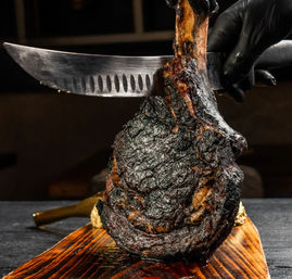 Close-up of a juicy tomahawk ribeye with deep charred crust being sliced from the bone with a large knife by gloved hands on a rustic wooden cutting board — smoky grilled steak presentation.