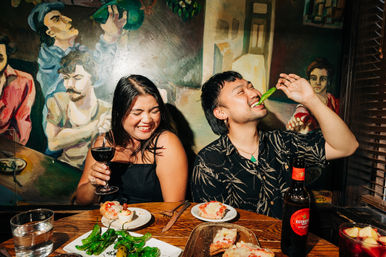 Two people laugh in a mural-lined tapas bar as one bites a blistered shishito pepper and the other sips red wine, with toasted bread, plates of peppers and a beer bottle on the wooden table.