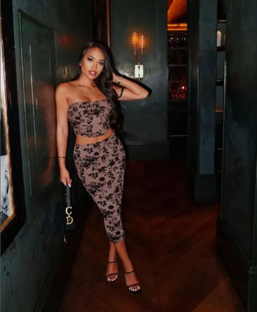 Stylish woman with long dark hair in a strapless floral-print two-piece (tube top and midi skirt) and strappy heels posing with a small black clutch in a dimly lit upscale lounge hallway.