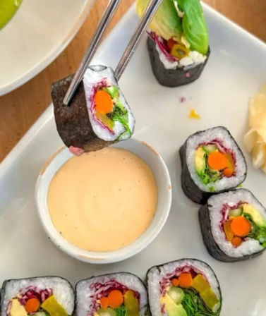 Close-up of colorful vegetarian sushi rolls wrapped in nori with rice, carrot, avocado, leafy greens and pickled vegetables — one held by chopsticks above a bowl of creamy spicy mayo on a white plate