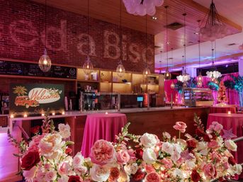 Pink-lit indoor event bar with exposed brick wall, hanging pendant lights, a 'Welcome' sign, round tables draped in pink linens and abundant pink and red rose arrangements.