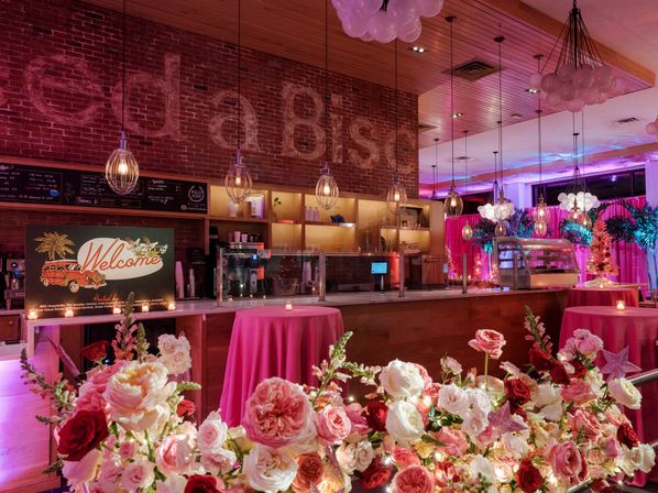 Pink-lit indoor event bar with exposed brick wall, hanging pendant lights, a 'Welcome' sign, round tables draped in pink linens and abundant pink and red rose arrangements.