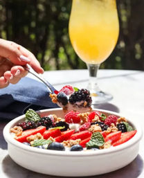 Hand with spoon scooping a yogurt and granola breakfast bowl topped with sliced strawberries, blueberries, blackberries, raspberries and mint, with a glass of orange juice on a sunny patio.