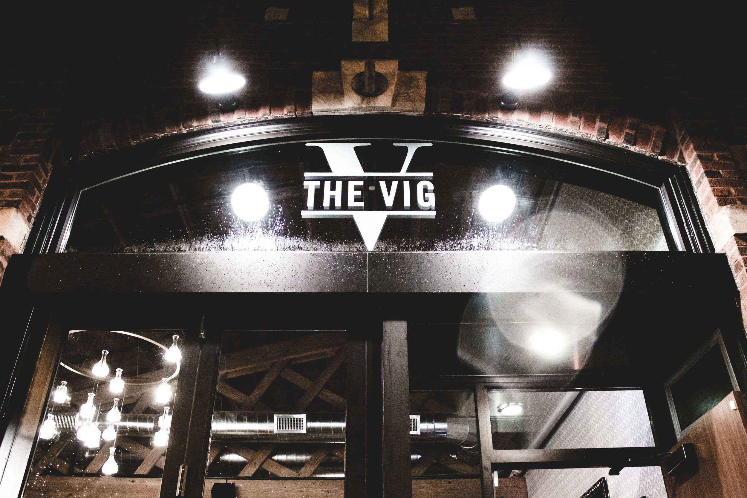 Moody nighttime urban brick storefront with arched glass entrance, wet glass reflections, glowing round lights and a modern chandelier inside.