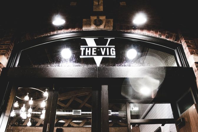 Moody nighttime urban brick storefront with arched glass entrance, wet glass reflections, glowing round lights and a modern chandelier inside.