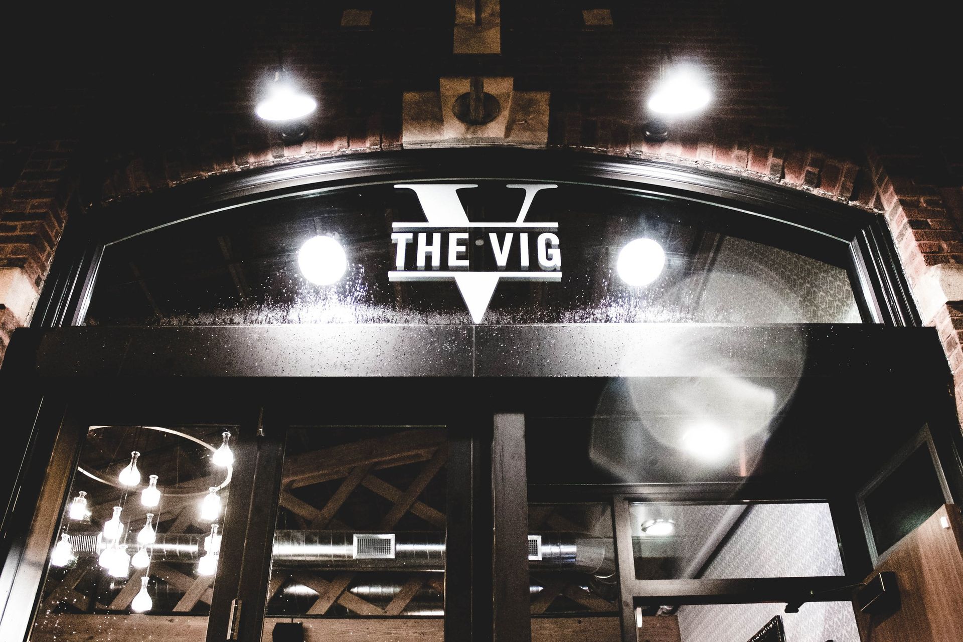 Moody nighttime urban brick storefront with arched glass entrance, wet glass reflections, glowing round lights and a modern chandelier inside.