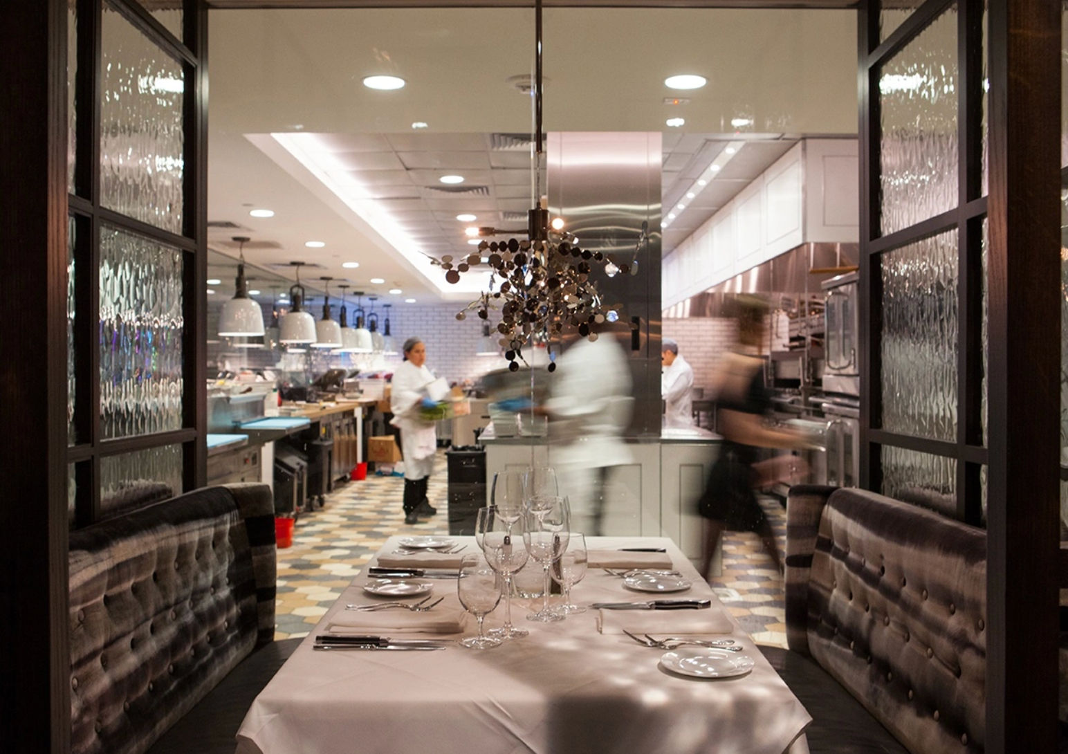 Fine-dining restaurant table set for four with white tablecloth, wine glasses and cutlery, overlooking a bustling open kitchen with chefs and blurred servers under pendant lights.