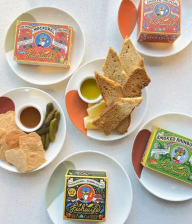 Flat-lay appetizer spread with colorful canned fish tins, toasted bread slices, round crackers, cornichons, lemon wedges and small bowls of olive oil and dipping sauce on white plates