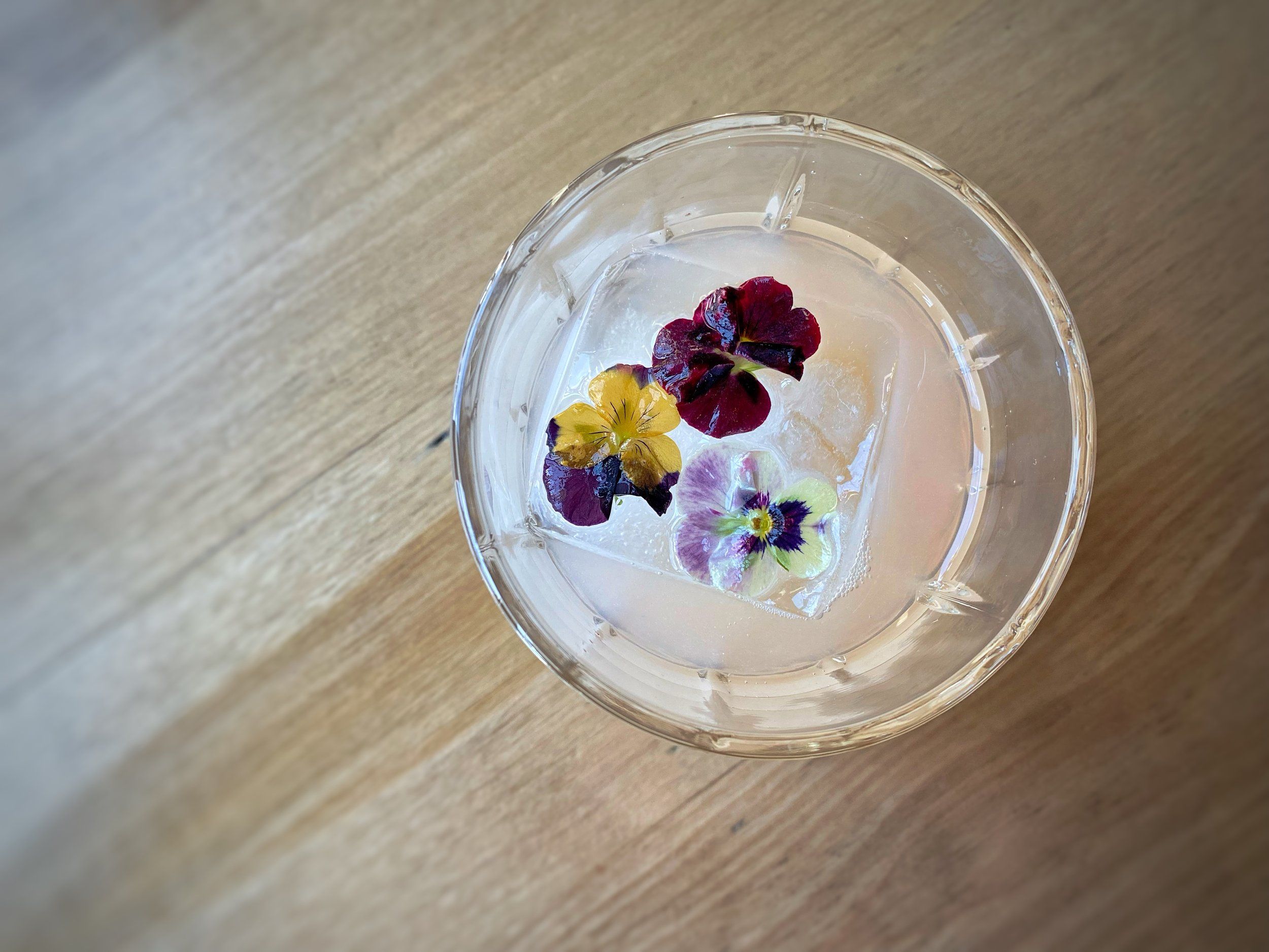 Top-down view of a pale pink cocktail in a cut-glass tumbler with a large ice cube topped by three colorful edible pansy flowers, resting on a light wood table.