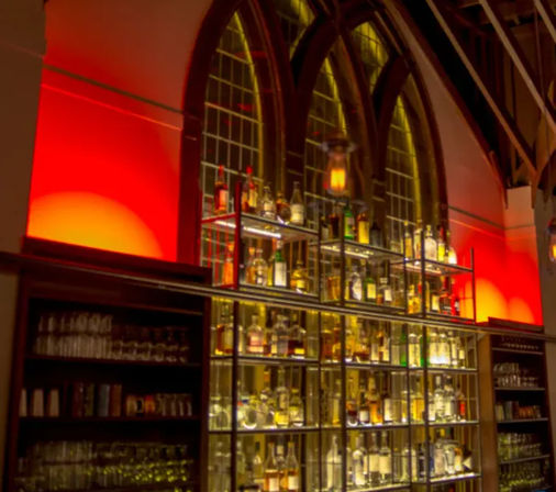 Interior of a dimly lit bar with tall arched windows, metal shelves of illuminated liquor bottles and glassware against warm red-orange accent lighting
