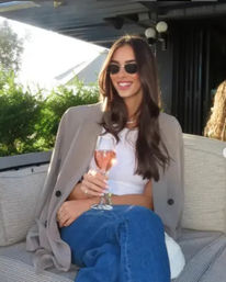 Smiling woman with long dark hair and sunglasses, blazer draped over her shoulders, white crop top and jeans, holding a glass of rosé while seated on an outdoor rooftop terrace lounge with greenery in the background.