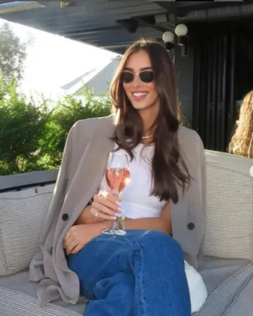 Smiling woman with long dark hair and sunglasses, blazer draped over her shoulders, white crop top and jeans, holding a glass of rosé while seated on an outdoor rooftop terrace lounge with greenery in the background.