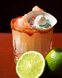 Chili-rimmed frozen lime cocktail in a cut-glass tumbler topped with pink shaved ice and a tiny sombrero garnish, with fresh lime halves — Mexican-inspired refreshing drink.