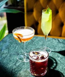 Trio of vibrant cocktails — orange coupe with citrus twist, pale green flute with leaf garnish, and red iced lowball — on a green marble table by a mustard tufted booth in a cozy bar