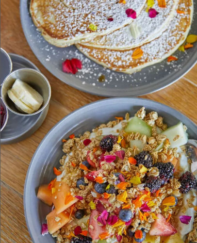 Colorful brunch spread on a wooden table: powdered-sugar pancakes with edible flower petals and a yogurt granola bowl topped with blackberries, blueberries, strawberries, melon and banana slices.