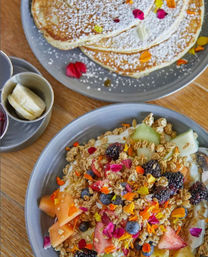 Colorful brunch spread on a wooden table: powdered-sugar pancakes with edible flower petals and a yogurt granola bowl topped with blackberries, blueberries, strawberries, melon and banana slices.