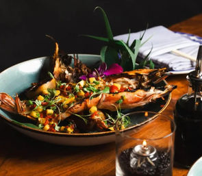 Grilled jumbo prawns topped with mango salsa and microgreens in a shallow ceramic bowl, garnished with an edible flower and tropical leaves on a wooden restaurant table beside a napkin, oil cruet and lit candle.
