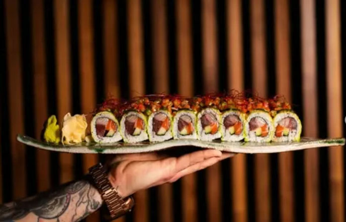 Tattooed hand holding a rectangular plate of green-wrapped sushi rolls topped with spicy red garnish, served with pickled ginger and wasabi against a wood-slat background.