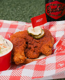 Crispy Southern-style spicy fried chicken sandwich on bread topped with pickle slices and a small red “HOT!” flag, served with a side of coleslaw on red checkered paper.