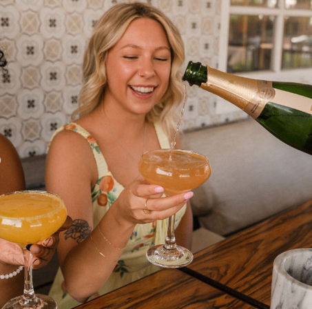 Smiling woman in a floral sundress at a brunch table holding a coupe glass as champagne is poured, with citrus cocktails and decorative tile wall in the background
