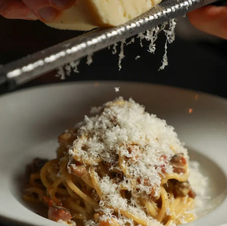 Close-up of a snowy shower of freshly grated Parmesan falling onto creamy spaghetti carbonara with pancetta in a white bowl